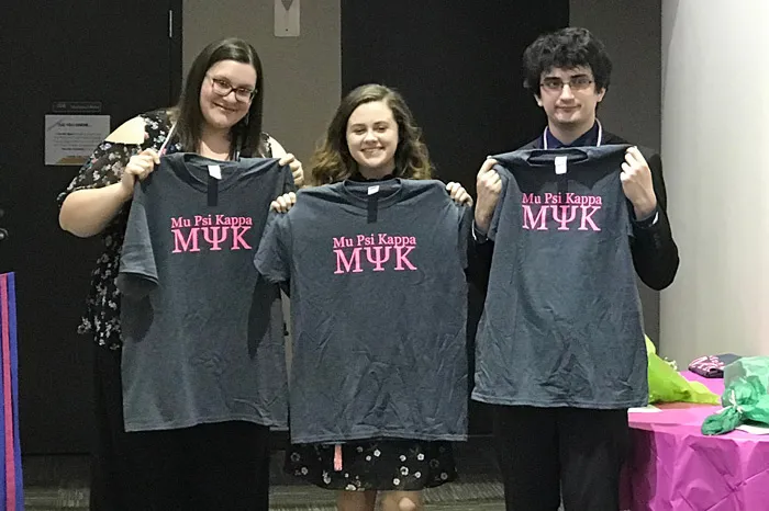 Three students holding up Mu Psi Kappa T-shirts