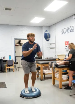 A Marietta College athlete attempts to catch a football while standing on a balance ball